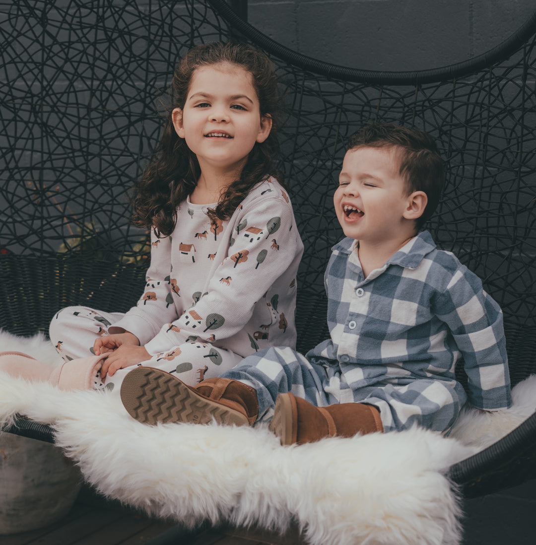 Two children in pajamas and sheepskin boots sitting on a white sheepskin rug in a chair