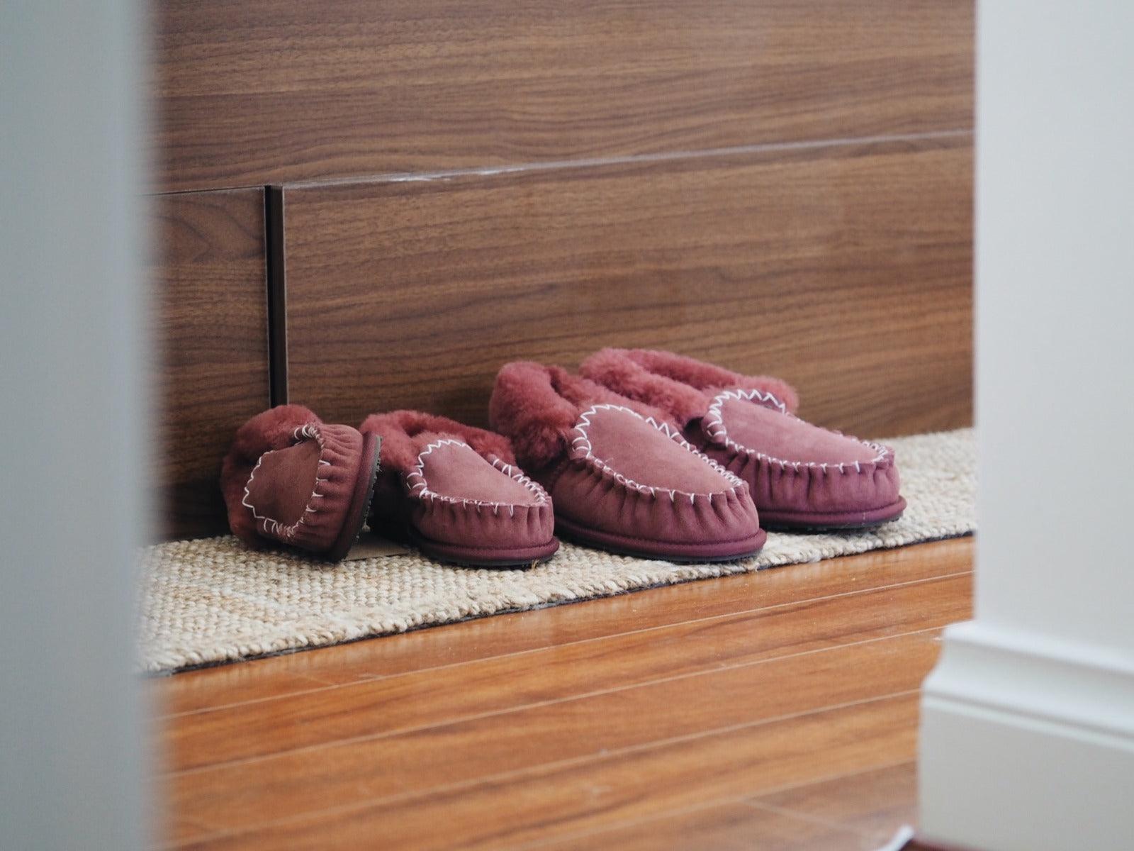 Pair of burgundy sheepskin moccasin slippers on a textured rug, wooden floor background