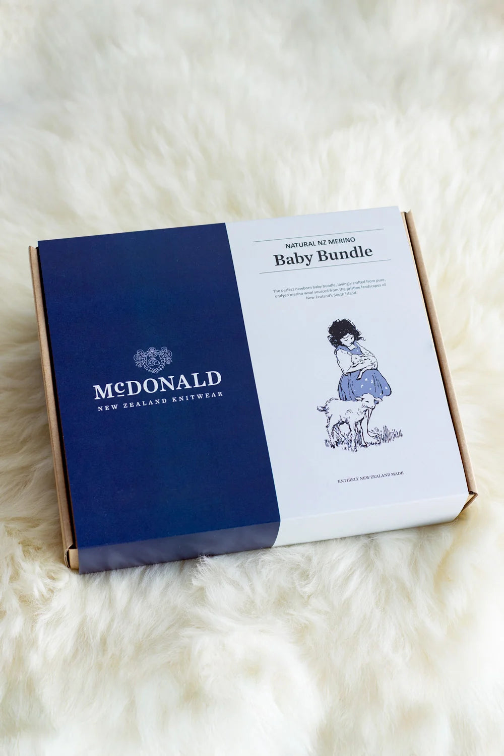 A Mcdonald New Zealand Baby Bundle box on a white faux fur background. The box is navy blue on the left with the Mcdonald logo, and white on the right with 'Baby Bundle' text and a vintage-style illustration of a mother and child - Newborn Baby Bundle — 100% Natural Merino Wool, Made in New Zealand