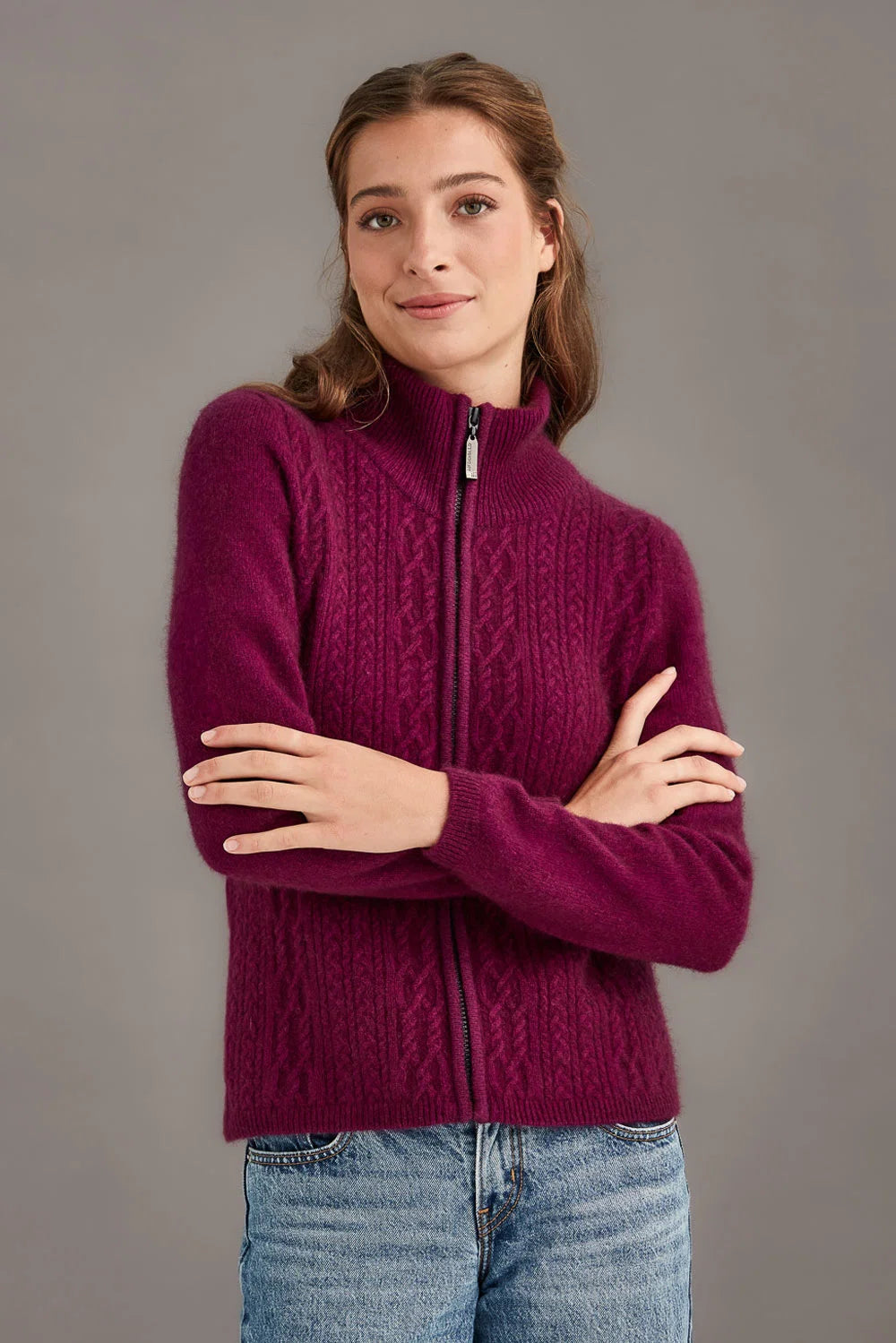 A woman wearing a deep magenta cable-knit zip-up sweater and blue jeans, standing against a solid gray studio background - Possum Merino Cable Zip Jacket — Made in New Zealand
