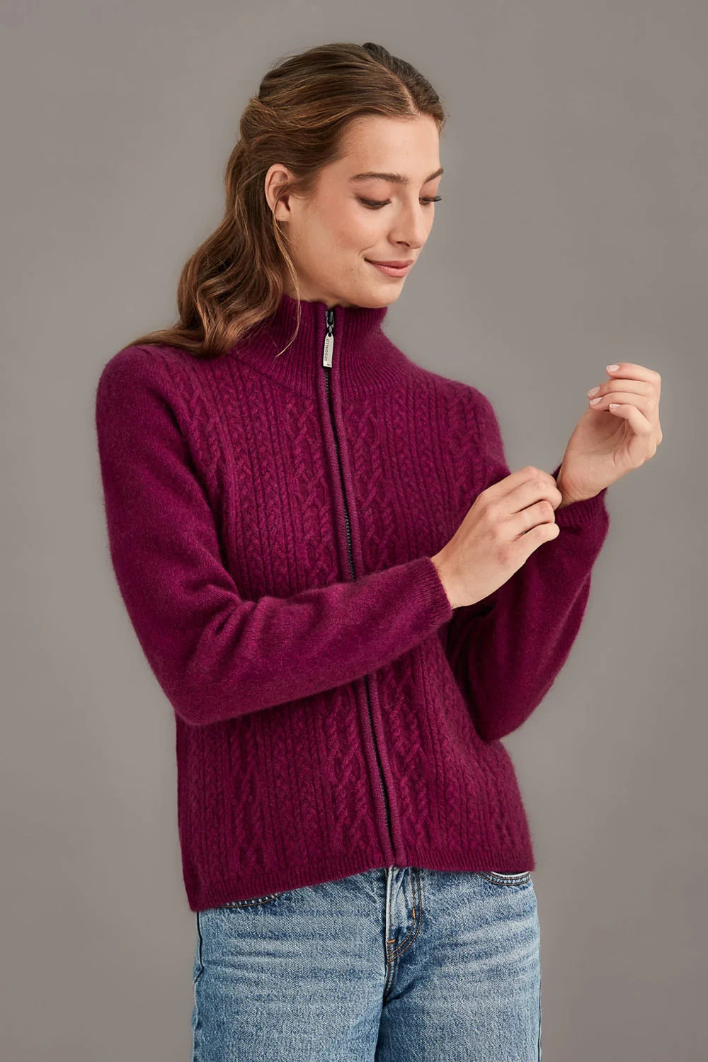 A woman wearing a deep magenta cable-knit zip-up sweater and blue jeans, standing against a solid gray studio background - Possum Merino Cable Zip Jacket — Made in New Zealand