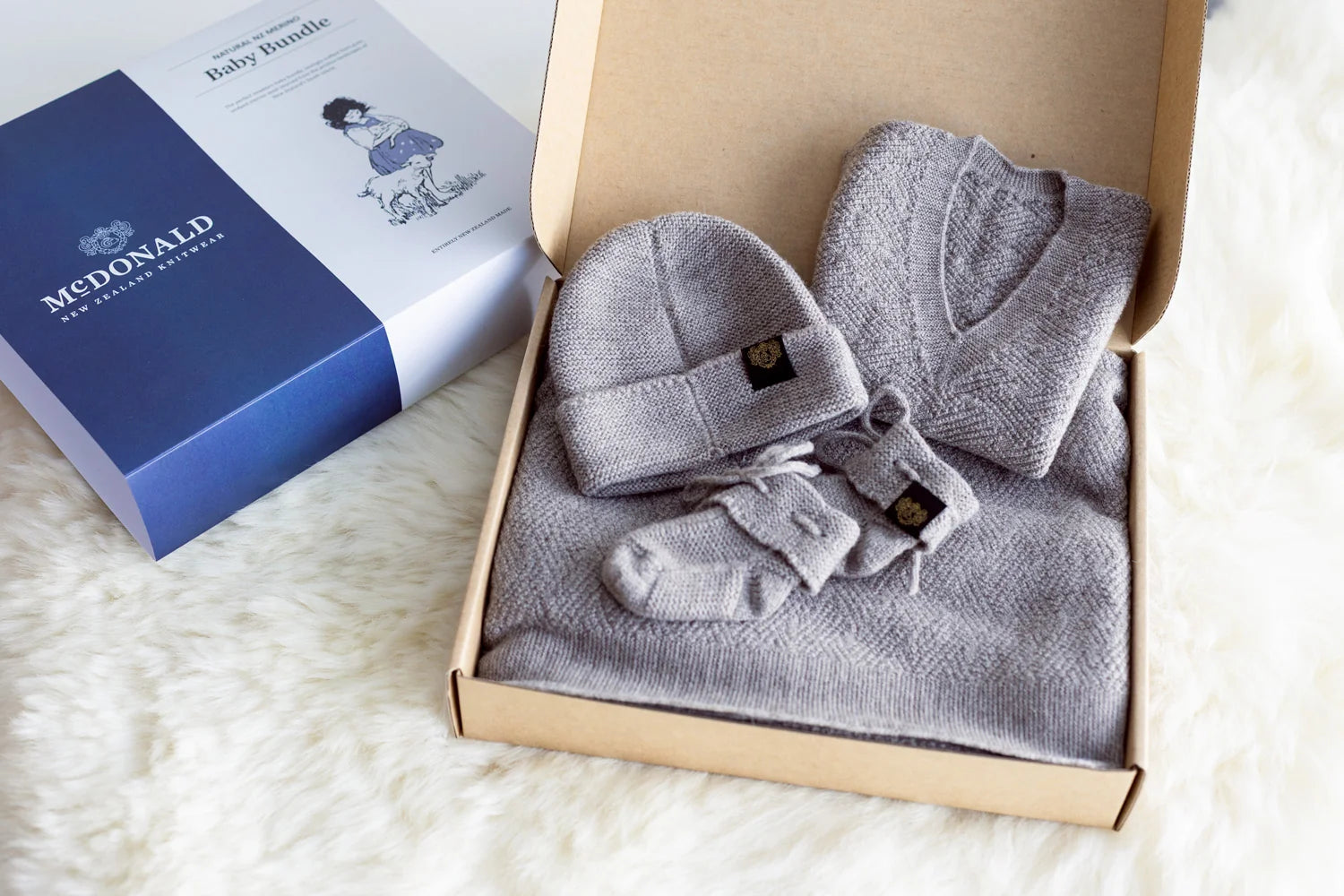 A baby gift set from McDonald's featuring a gray knit baby hat, booties, and sweater arranged in a kraft box next to its blue and white branded packaging on a white faux fur surface - Newborn Baby Bundle — 100% Natural Merino Wool, Made in New Zealand