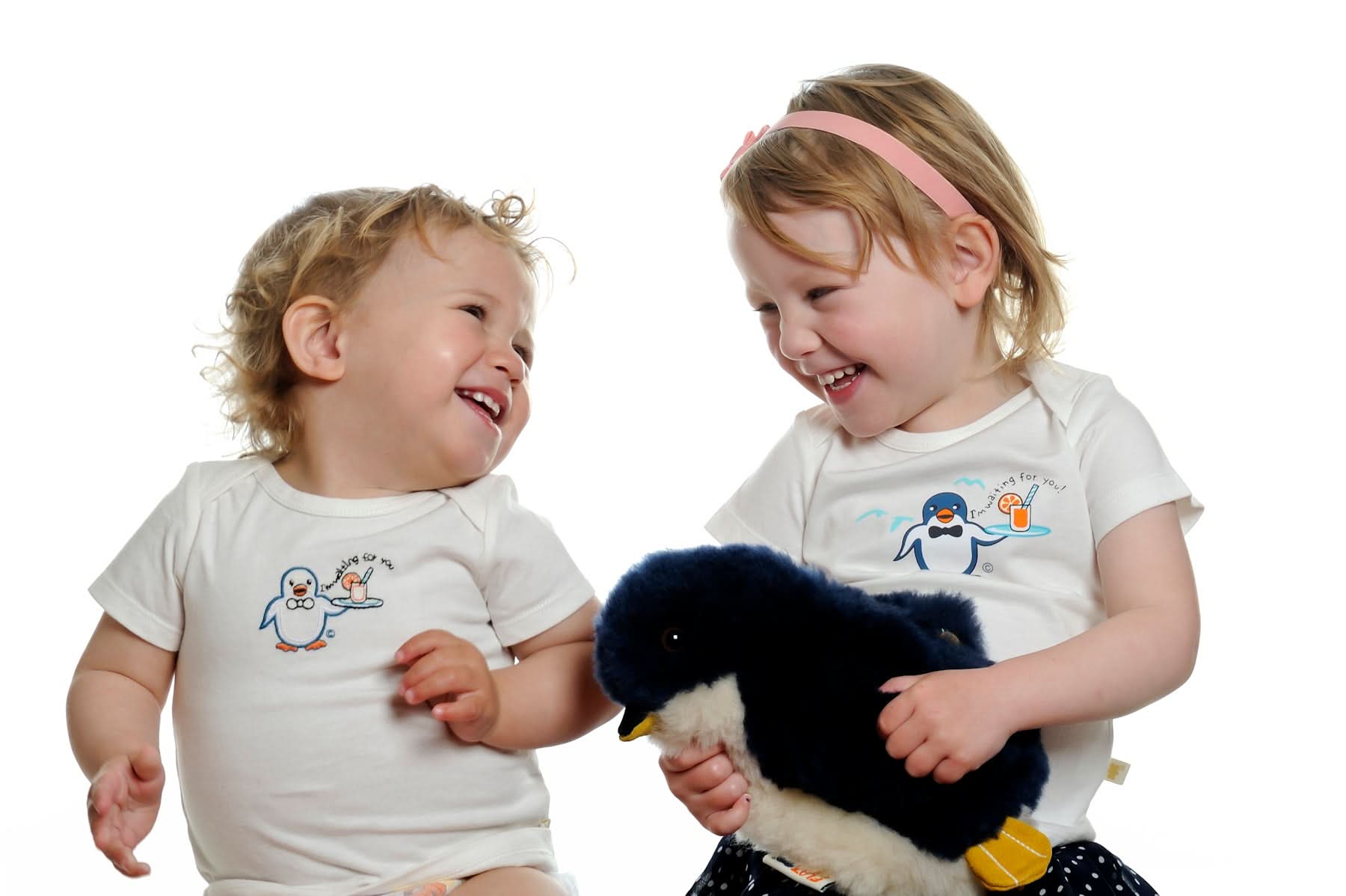 Two toddlers holding a soft black and white plush penguin toy against white background. - Yellow Earth Australia