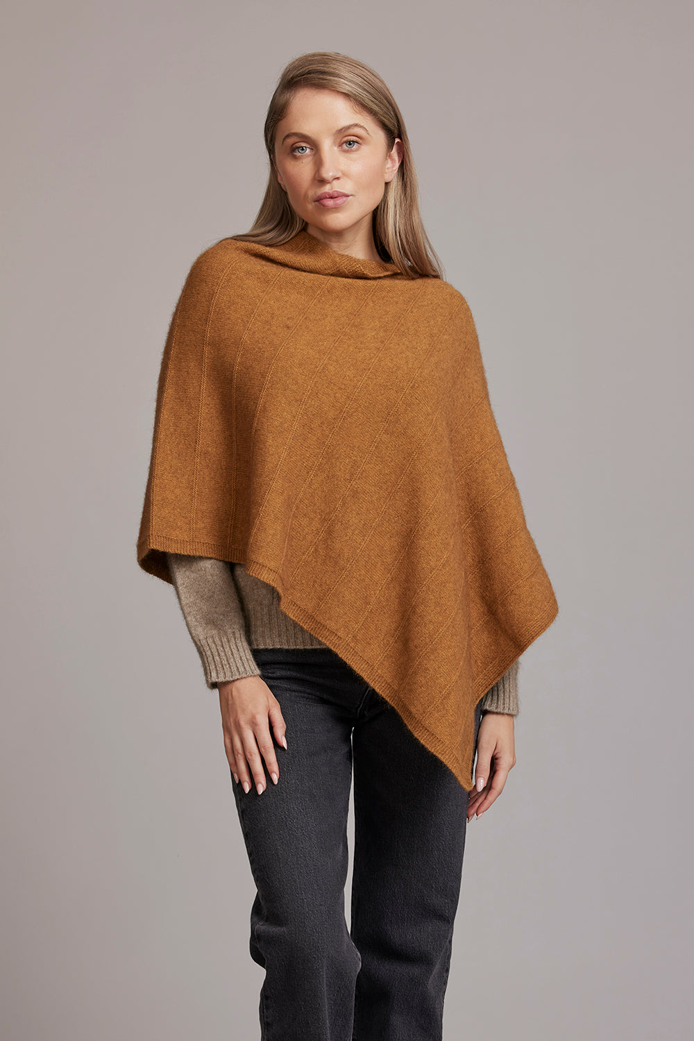 Woman wearing caramel wool poncho with ribbed sleeves over dark jeans, front view. - Yellow Earth Australia