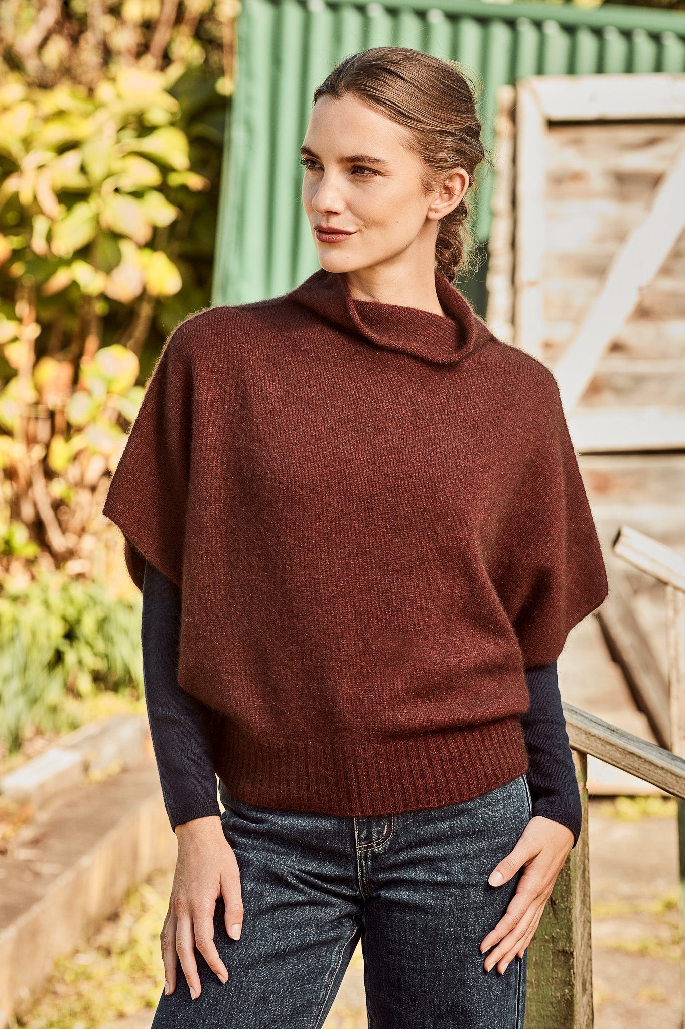 A woman wearing a dark brown cowl-neck poncho sweater - the Women's Shrug Possum Merino Wool Silk Sweater - over a black long-sleeve top and blue jeans, standing outdoors in front of a green corrugated metal wall and wooden structure.
