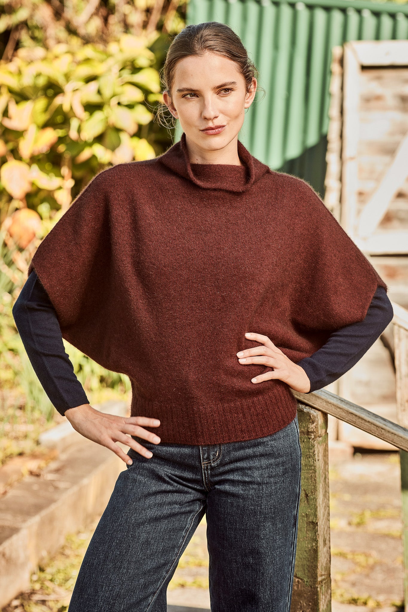 A woman wearing a dark brown ribbed knit poncho sweater - the Women's Shrug Possum Merino Wool Silk Sweater - over a navy blue long-sleeve shirt and dark blue jeans, posing outdoors against a rustic green and wood background.