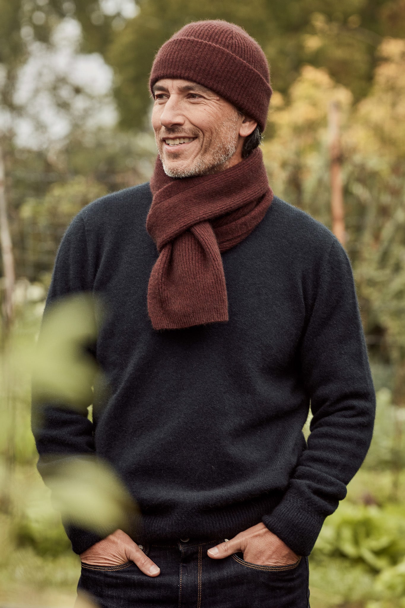 A smiling man outdoors wearing a dark charcoal knit sweater, a rust-brown knit beanie, and a matching rust-brown knit scarf, with a blurred natural background - with Possum Merino Wool Silk Fine Rib Beanie and Scarf in Waina colour