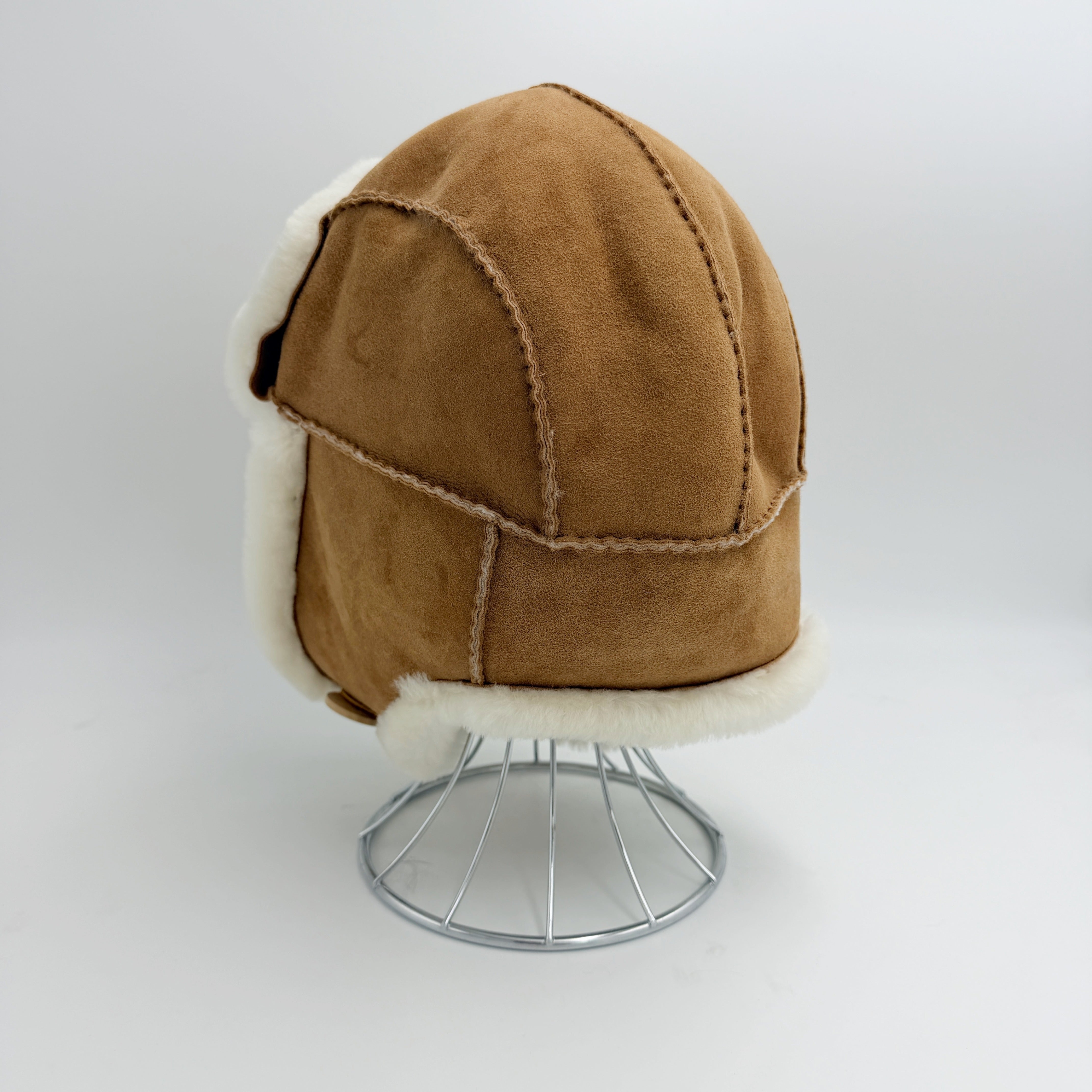 A tan suede aviator-style hat with white shearling lining, displayed on a silver wire stand against a light gray background - Yellow Earth