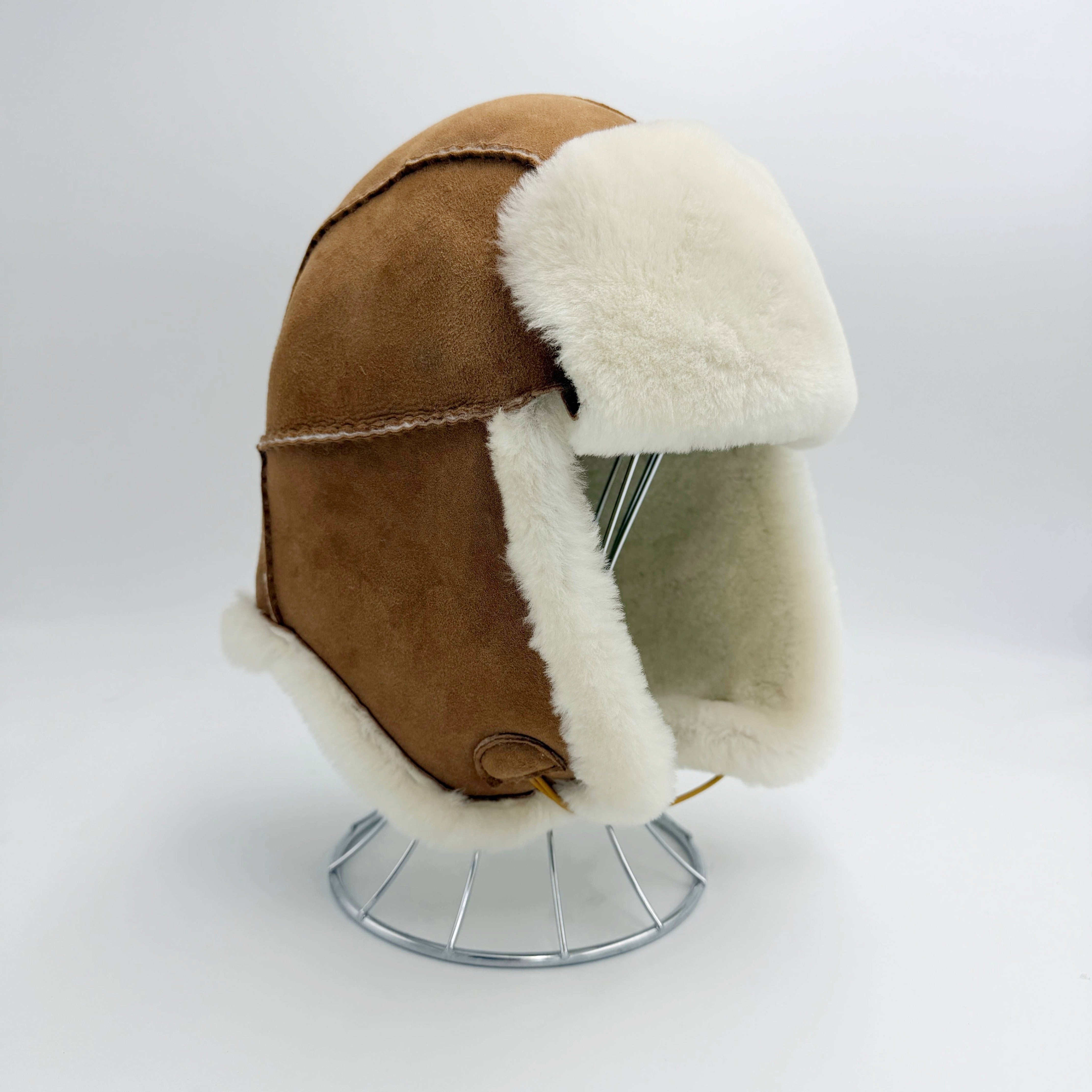 A brown suede aviator-style helmet with thick white shearling lining, displayed on a silver metal stand against a light gray studio background - Yellow Earth