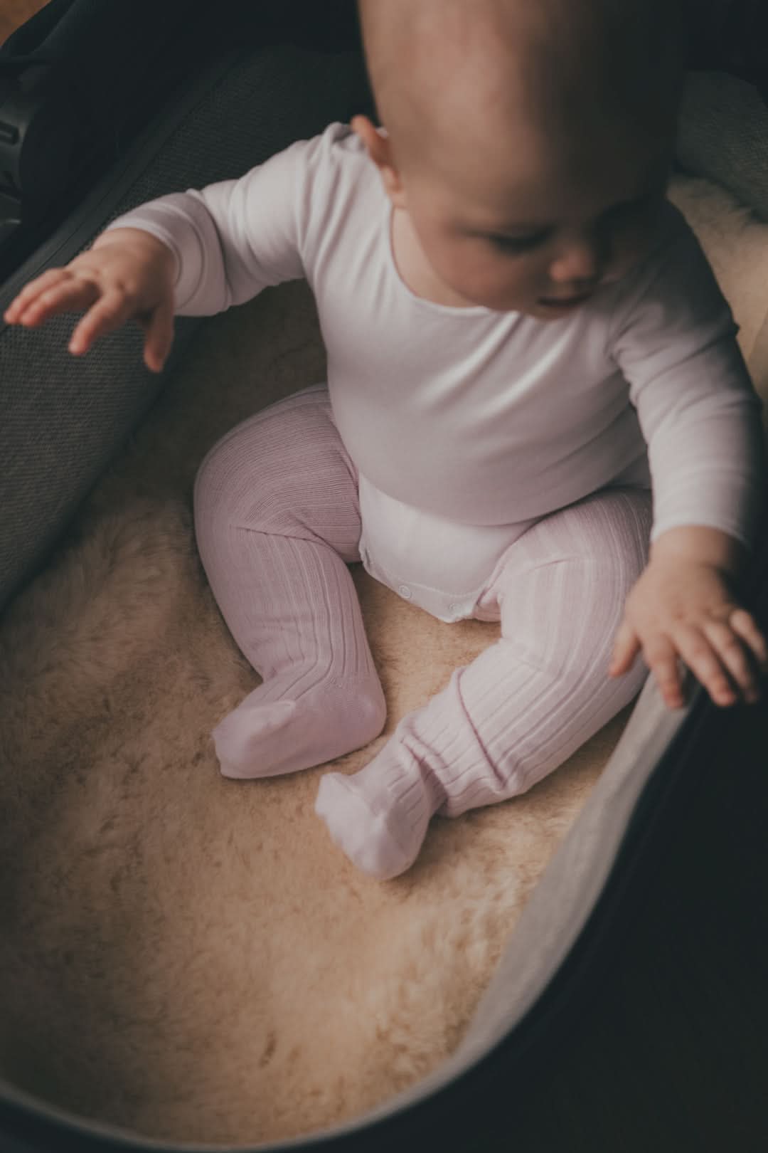 Baby sitting on natural sheepskin in pink ribbed knit outfit, overhead view. - Yellow Earth Australia